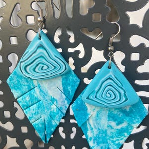 May include: A pair of dangle earrings featuring a triangular top piece with a teal and white swirl pattern, attached to a larger, leaf-shaped bottom piece with a mottled teal and white texture. The earrings hang from silver-toned metal hooks.
