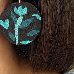 May include: A circular earring featuring a textured black surface with a painted light blue and dark teal floral design. The earring is attached to a square silver-toned stud with small embedded clear stones, creating a contrast between the dark base and the bright botanical pattern.