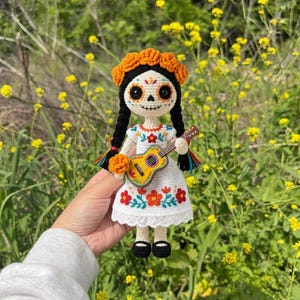 Op de afbeelding: Een gehaakte pop in een witte jurk met geborduurde oranje en rode bloemen, met een gele gitaar. De pop heeft zwarte vlechten met een oranje bloemenkrans en een wit gezicht met zwarte oogkassen en een gestikt neusdetail.