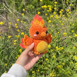 May include: A small, plush, crocheted phoenix toy featuring vibrant orange and red yarn, a textured crest on its head, two black bead eyes, and a soft yellow belly. The creature has small wings and a rounded body, crafted with soft, fuzzy yarn.