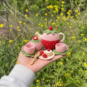 Può includere: Un set da tè in miniatura all'uncinetto composto da una teiera a forma di fragola con coperchio a foglia verde, una zuccheriera abbinata, due tazze rosa su piattini a foglia, un piattino con una tortina e due fragole, e un cucchiaino di legno.