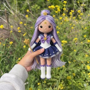 May include: A crocheted doll with long lavender hair styled in a top knot, wearing a white and dark blue sailor-style outfit with gold star accents, a purple bow, and white boots. The doll holds a small gold star-topped wand in its hand.