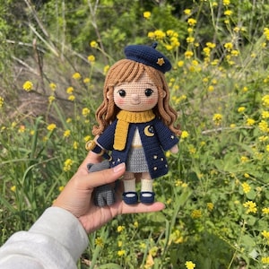 May include: A crochet doll with wavy brown hair wearing a dark blue beret, a matching coat with yellow star embroidery, a yellow scarf, a gray skirt, and dark blue shoes. The doll holds a small telescope and stands next to a tiny gray cat figure.