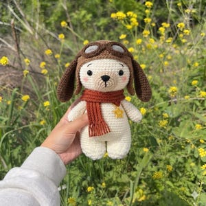 Op de afbeelding: Een crèmekleurig gehaakt knuffeldier met lange bruine oren, voorzien van een bruine pilotenmuts met een bril, een roestoranje sjaal en een kleine gele sterbedel. Het speelgoed heeft zwarte ogen, een zwarte geborduurde neus en roze blosjes.