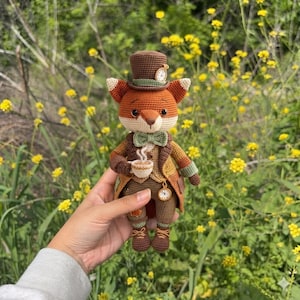 Puede incluir: Una figura de zorro tejida a ganchillo que lleva un sombrero de copa marrón con un pequeño reloj, una pajarita verde, un chaleco beige y una chaqueta marrón. El zorro sostiene una pequeña taza blanca con vapor y tiene un reloj redondo en el chaleco.