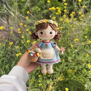 May include: A crocheted doll with brown hair in pigtails, wearing a blue dress with a white apron and a yellow and white flower crown. The doll holds a small brown basket filled with colorful flowers and has a tiny butterfly on its shoulder.