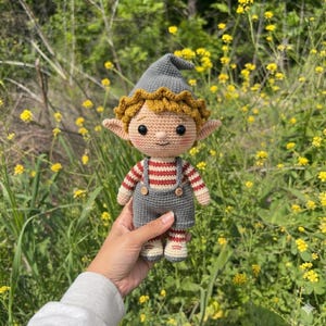 May include: A crocheted doll with a pointed gray hat, a mustard yellow ruffled brim, and large black eyes. The doll wears a red and white striped long-sleeve shirt under gray overalls with two small wooden buttons on the front straps and matching shoes.