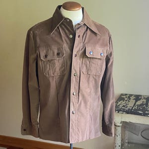 May include: A brown corduroy shirt with a button-down front, two chest pockets, and long sleeves. The shirt has a classic collar and buttoned cuffs. The buttons are a mix of brown and blue. The shirt is displayed on a mannequin.