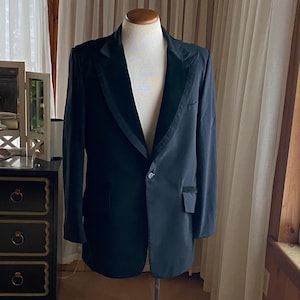 May include: A dark blue blazer with a black velvet lapel and a single button closure. The blazer is displayed on a mannequin. The blazer has a pocket on the left side. The background includes a window and a dark dresser.