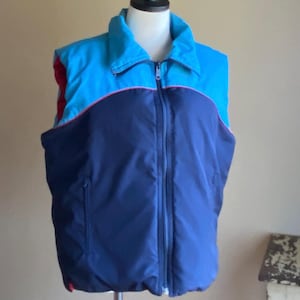 May include: A blue, red, and pink sleeveless puffer vest with a zipper closure. The vest has a stand-up collar and a zippered pocket on the left side.