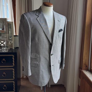 May include: A black and white gingham blazer displayed on a mannequin. The jacket features a notched lapel, a three-button front closure, two flap pockets at the waist, and a small dark pocket square tucked into the chest pocket.