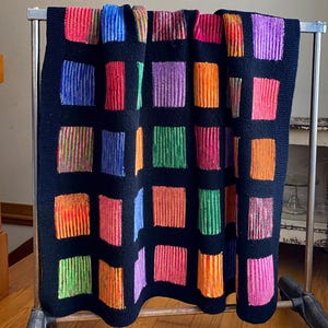May include: A black knitted blanket with a pattern of colorful, vertical striped squares. The squares are in shades of red, orange, yellow, green, blue, and purple. The blanket is displayed on a metal rack.