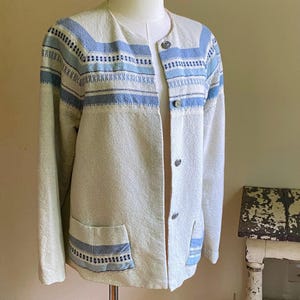 May include: A white jacket with blue and white striped trim. The jacket has a button closure and two pockets.