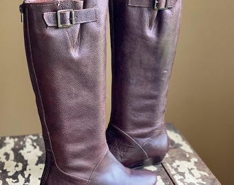 May include: A pair of brown leather knee-high boots with a buckle detail on the top of the shaft. The boots have a slight wedge heel.