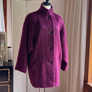 May include: A deep magenta, fuzzy-textured coat featuring a high collar, long sleeves, and a front button closure with four dark buttons. The coat includes two slanted welt pockets on the front and has a loose, straight-cut silhouette.