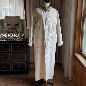 May include: A long, off-white duster coat featuring a tan corduroy collar, long sleeves, and a front button-down closure. The garment includes two chest pockets and two lower hip pockets, all with flaps, and is displayed on a black mannequin.