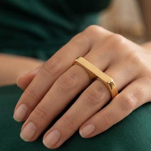 Puede incluir: Un anillo doble de tono dorado con un diseño de barra rectangular lisa que se extiende sobre dos dedos. El anillo presenta una superficie pulida y reflectante, con una estructura de banda metálica continua y bordes redondeados y definidos.