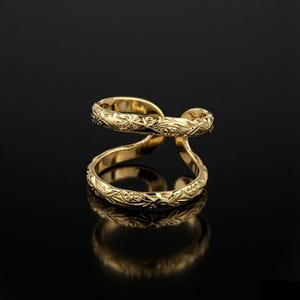 May include: A gold-toned ring featuring a double-band design with an open gap. The surface of the metal is engraved with a continuous, intricate floral or leaf-like pattern that wraps around both bands, creating a textured and reflective finish.