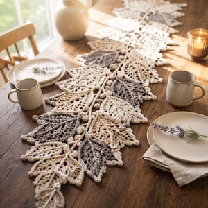 May include: A long, crocheted table runner featuring a repeating pattern of interconnected leaves in shades of cream, beige, and charcoal gray. The design includes small, round, crocheted berry-like accents along the central vine structure.