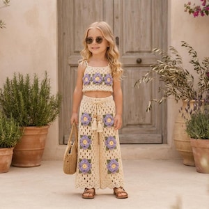 Puede incluir: Un conjunto de dos piezas de ganchillo compuesto por un top corto y pantalones anchos de color crema. Ambas piezas presentan motivos cuadrados con centros florales morados y amarillos y hojas verdes. El pantalón tiene un cordón con borlas.