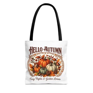 Può includere: Una borsa tote bianca con manici neri caratterizzata da una grafica di zucche, funghi e foglie autunnali. Il testo HELLO AUTUMN è stampato in marrone sopra l'illustrazione, e il testo Cozy Nights & Golden Leaves è stampato sotto.