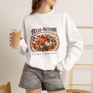 Può includere: Una felpa bianca a maniche lunghe con una stampa grafica di zucche, funghi e foglie autunnali. Il testo recita HELLO AUTUMN e Cozy Nights & Golden Leaves. La felpa è abbinata a pantaloncini di jeans grigi e un bicchiere trasparente.