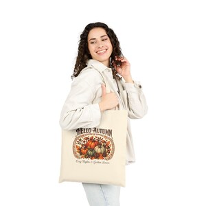 Può includere: Una borsa tote in tela color crema con una stampa grafica di zucche, funghi e foglie autunnali. Il testo "HELLO AUTUMN" è riportato in alto e "Cozy Nights & Golden Leaves" in basso, incorniciato da un bordo ovale decorativo.