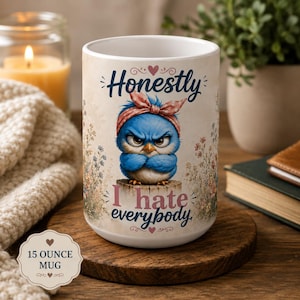 Op de afbeelding: Een witte 15-ounce keramische mok met een afbeelding van een chagrijnige blauwe vogel met een roze hoofdband met stippen, zittend op een boomstronk. De tekst luidt Honestly I hate everybody, omringd door kleine bloemillustraties en hartjes.