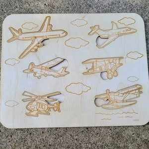 May include: Wooden puzzle board with six cutouts of different types of aircraft, including a jet, a propeller plane, a biplane, a helicopter, and a seaplane. The board is made of light wood and has a natural finish.