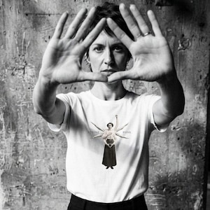 Może przedstawiać: Biały t-shirt z krótkim rękawem z centralną grafiką przedstawiającą postać z wieloma ramionami ułożonymi w wachlarz. Napis STRIKE POSE znajduje się po obu stronach postaci. Koszulka jest zestawiona z gładkimi czarnymi spodniami.