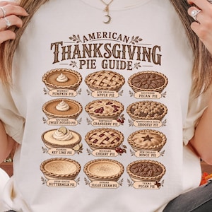 Puede incluir: Una camiseta de manga corta color crema con un gráfico marrón titulado American Thanksgiving Pie Guide. El diseño muestra doce ilustraciones de varios pasteles con etiquetas que incluyen Pumpkin, Apple, Pecan, Sweet Potato y Cranberry.
