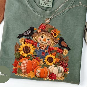 Peut inclure: Un t-shirt vert mousse avec un motif représentant un épouvantail portant un chapeau en patchwork, entouré de deux oiseaux noirs, de tournesols et de trois citrouilles. Le t-shirt porte une étiquette Comfort Colors et un collier argenté.