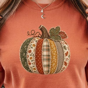 May include: A burnt orange short-sleeve t-shirt featuring a central graphic of a pumpkin composed of various fabric-like patterns in shades of green, yellow, brown, and cream, with a dark green stem and leaf detail at the top of the pumpkin.