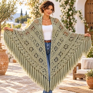 May include: A sage green triangular crochet shawl featuring an intricate floral pattern with openwork details and long, flowing fringe along the bottom edges. The garment is worn over a white top and blue jeans, showcasing the detailed needlework.