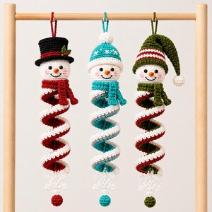 May include: Three crocheted snowman ornaments hanging from a wooden rod. Each snowman has a white head, a colorful scarf, and a long, spiraling body in red, teal, or green. They feature hats, matching scarves, and white snowflake accents.