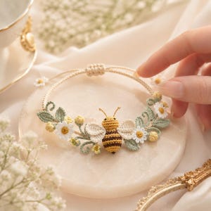 Könnte beinhalten: Ein zartes Häkelarmband mit einem zentralen Bienenmotiv mit braunen und gelben Streifen, flankiert von weißen Gänseblümchen, kleinen gelben Knospen und salbeigrünen Blättern. Das Armband hat eine dünne, verstellbare Kordel mit Schiebeknoten.