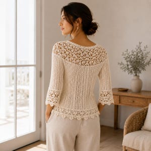 May include: A cream-colored, three-quarter sleeve top featuring an intricate crochet yoke, vertical ribbed knit detailing on the torso, and a scalloped floral crochet hemline. The garment has a wide neckline and a semi-sheer, lightweight texture.