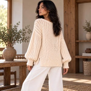 Peut inclure: Un pull en tricot épais de couleur crème avec une encolure en V, des manches longues bouffantes à poignets côtelés et des fentes latérales à l'ourlet, associé à un pantalon blanc ample. Le pull présente une texture côtelée verticale.