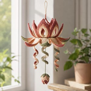 Pode incluir: Um ornamento de crochê suspenso em forma de flor de lótus, com pétalas em camadas em tons de rosa seco, creme e verde sálvia. Três gavinhas em espiral descem do centro, terminando num pequeno botão fechado na parte inferior da peça.
