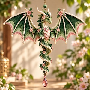 Könnte beinhalten: Ein hängendes Häkel-Drachenornament mit dunkelgrünen und cremefarbenen Flügeln und rosa Blumenakzenten. Der Drache hat einen gewundenen Körper, cremefarbene Hörner und grüne Schuppen, die mit kleinen rosa Blüten und Blättern verziert sind.