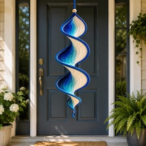 May include: A spiral-shaped hanging decoration featuring a crochet design with a color gradient transitioning from deep blue to light blue and cream. The object is suspended by a string attached to a small wooden bead at the top, creating a vertical twist.