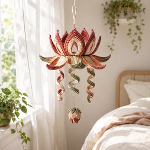 May include: A hanging crochet lotus flower mobile featuring layered petals in shades of terracotta, cream, and sage green. Three spiraling tendrils extend downward from the center, ending in a small, closed lotus bud at the bottom of the structure.