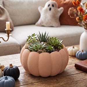 Peut inclure: Un pot de fleurs en forme de citrouille de couleur pêche mate rempli de diverses plantes succulentes vertes. Le pot repose sur une surface en bois avec un candélabre en laiton à quatre bougies crème, des citrouilles décoratives et un mug à tête de mort.