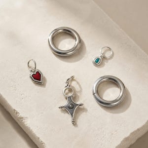 Könnte beinhalten: Eine Sammlung silberfarbener Schmuckkomponenten, bestehend aus zwei schlichten kreisförmigen Ringen, einem kleinen Anhänger mit roter Herzeinlage, einem kleinen Anhänger mit türkisfarbenem Stein und einem kreuzförmigen Anhänger.