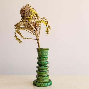 May include: A green ceramic vase with a textured, stacked design holds a dried Banksia flower. The vase has a glossy finish and a unique, handcrafted appearance. The flower has a brown, textured center and yellow-green accents.