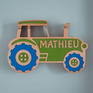 Puede incluir: Una decoración de pared de madera con forma de tractor, con un marco de madera clara, detalles de fieltro verde en el techo y la carrocería, y ruedas de fieltro azul. El nombre MATHIEU está recortado en letras mayúsculas en la sección de fieltro verde.