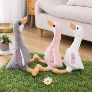 Peut inclure: Trois oies en peluche de couleur grise, rose et blanche, chacune dotée d'un bec jaune et du mot SCOTTIE brodé en rose sur l'aile. Ces animaux en peluche ont un long cou, des yeux noirs et des pattes jaunes, posés sur une surface verte.