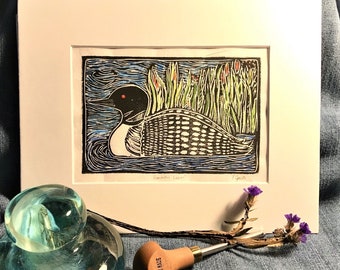 May include: A black and white woodblock print of a common loon swimming in a lake with green reeds. The print is framed in a white mat and has the text "Common Loon" and "Piegat" written below the image.