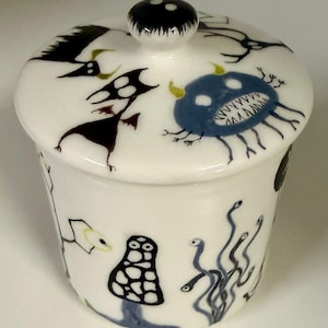 Può includere: Un barattolo in ceramica bianca con coperchio, caratterizzato da illustrazioni dipinte a mano di varie piccole creature e forme astratte in nero, blu e giallo. Il coperchio ha un pomello centrale e il corpo è cilindrico con finitura lucida.