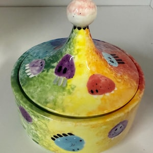 May include: A round ceramic lidded jar featuring a vibrant rainbow-colored gradient background. The surface is decorated with small, hand-painted colorful creatures with black dots for eyes and short legs, topped with a spherical knob handle.